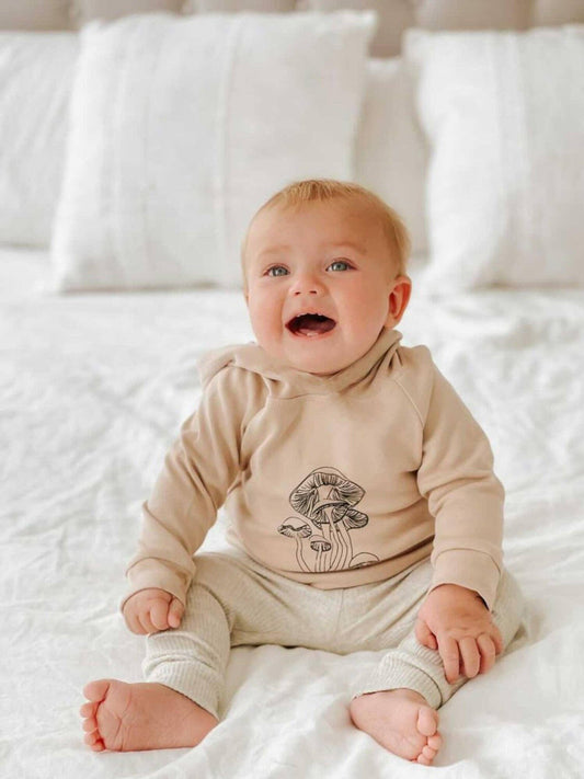 Baby wearing a beige hoodie with a mushroom design on it sitting on a white bed