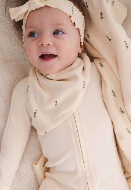 Baby wrapped in a beige swaddle with a matching headband and bib, lying on a soft surface.