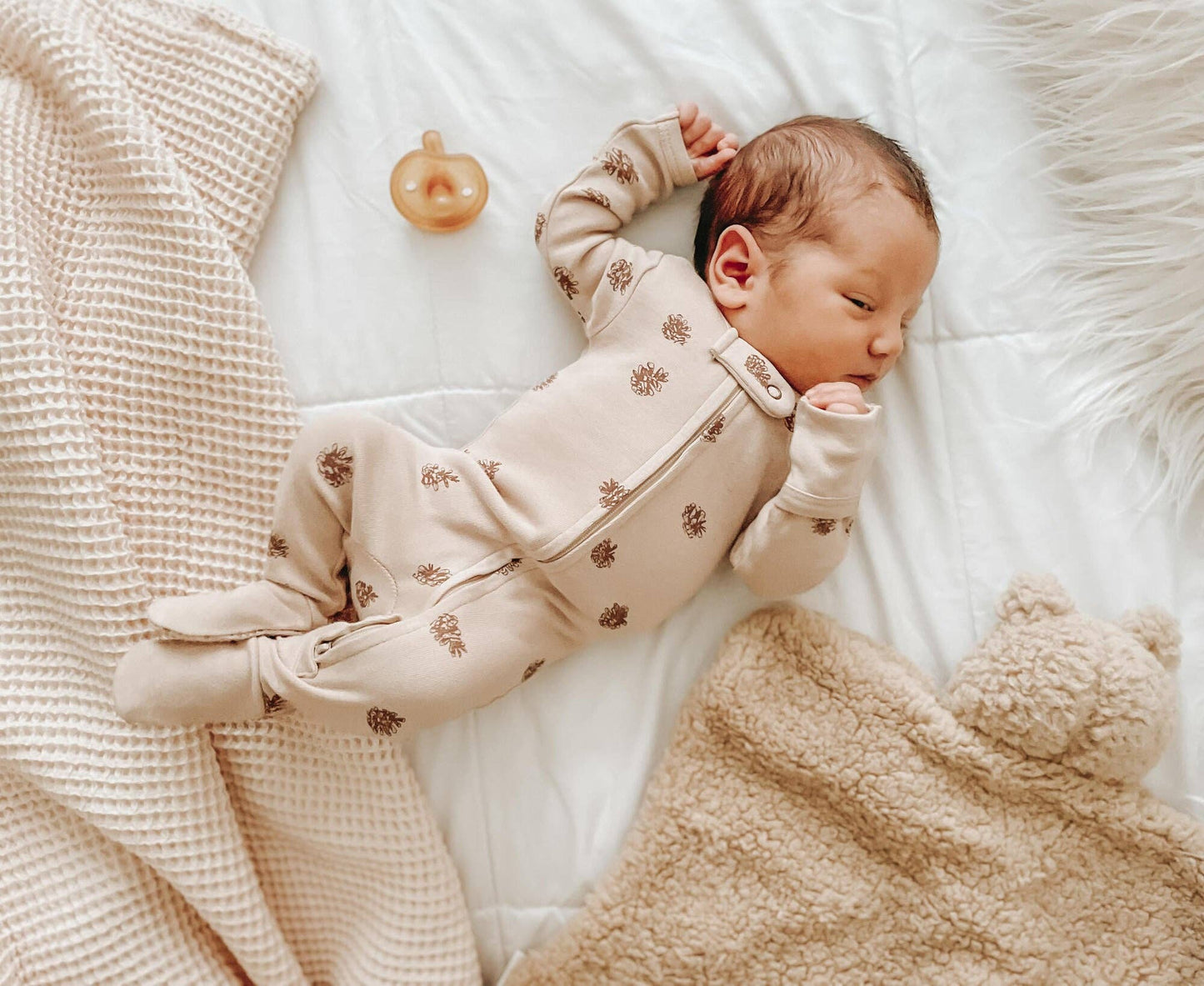 Newborn baby in a beige pinecones footie lying on a soft surface with a teddy bear lovey and a pacifier.