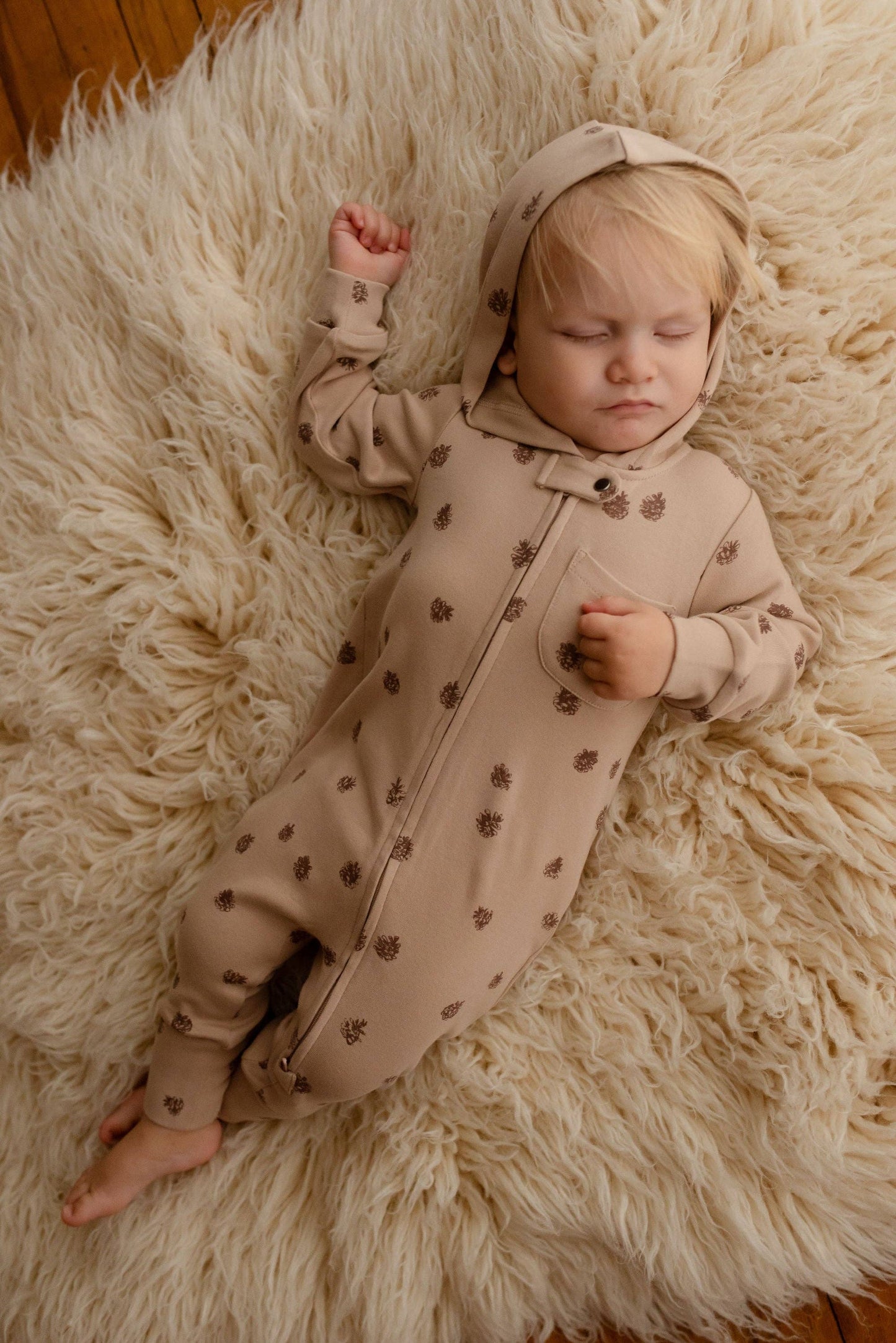 Baby in a beige romper with brown pinecones print lying on a fluffy white rug.