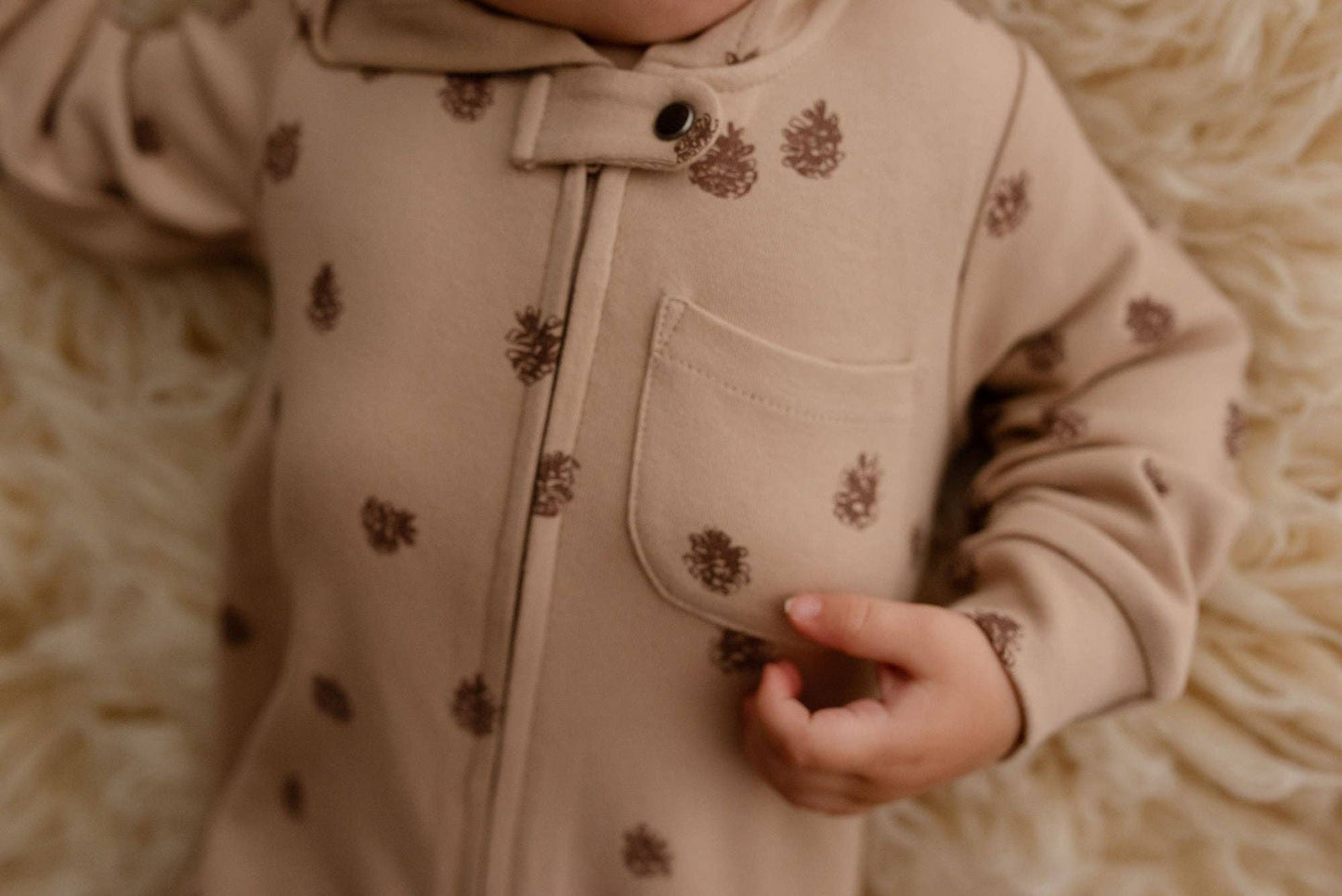 Close-up of a pocket on a beige hooded romper with pinecone patterns on a soft, beige background