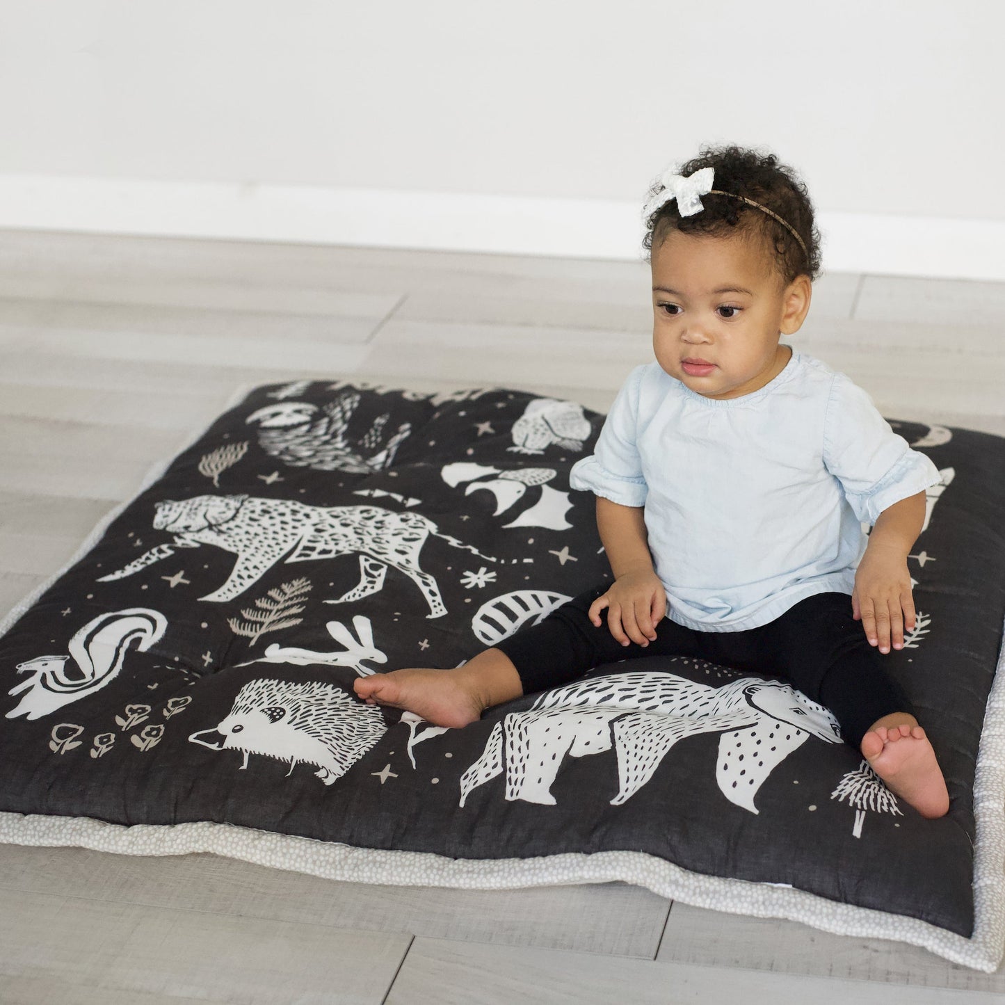 Child sitting on a black blanket with white animal illustrations on a light wooden floor.