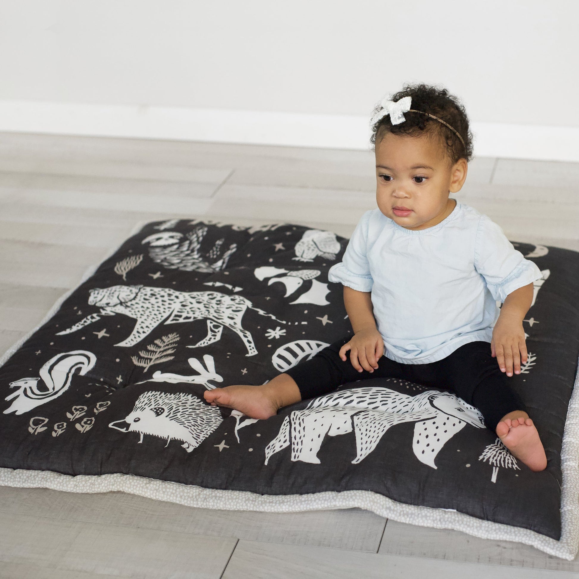 Child sitting on a black blanket with white animal illustrations on a light wooden floor.