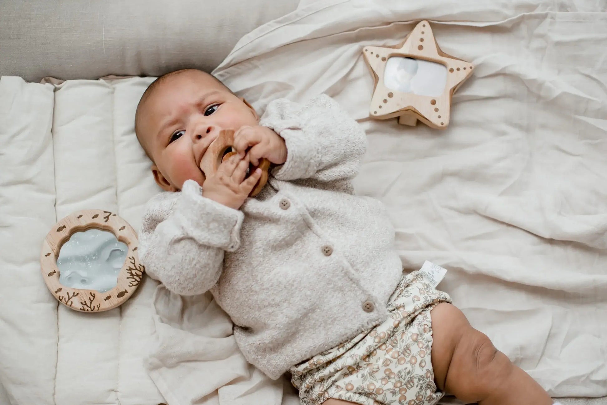 Baby lying on a soft bed teething on a wooden rattle with a wooden mirror and photo frame nearby