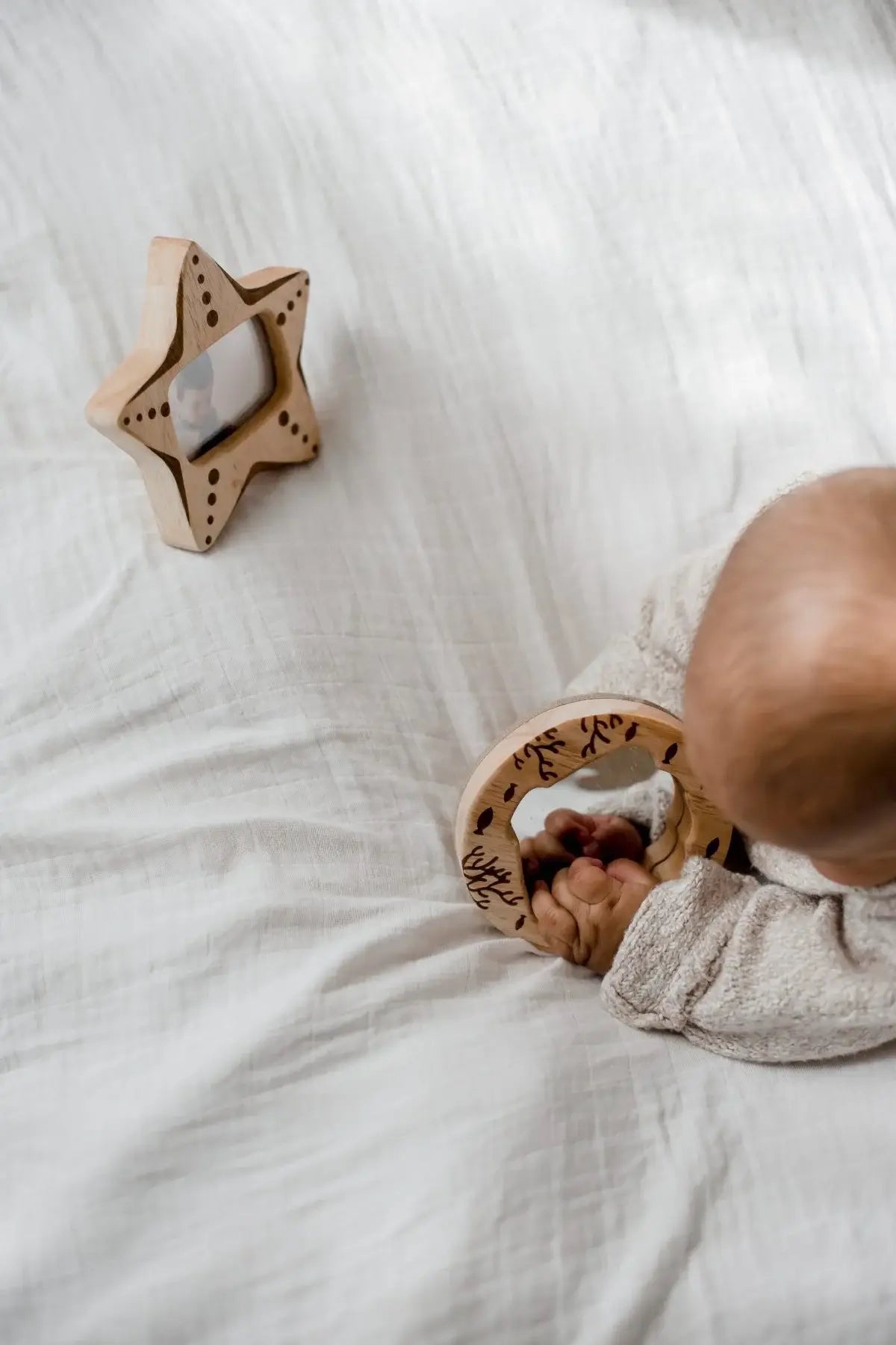 Baby laying on their stomach on a bed with wooden star and wooden mirror and rattle 