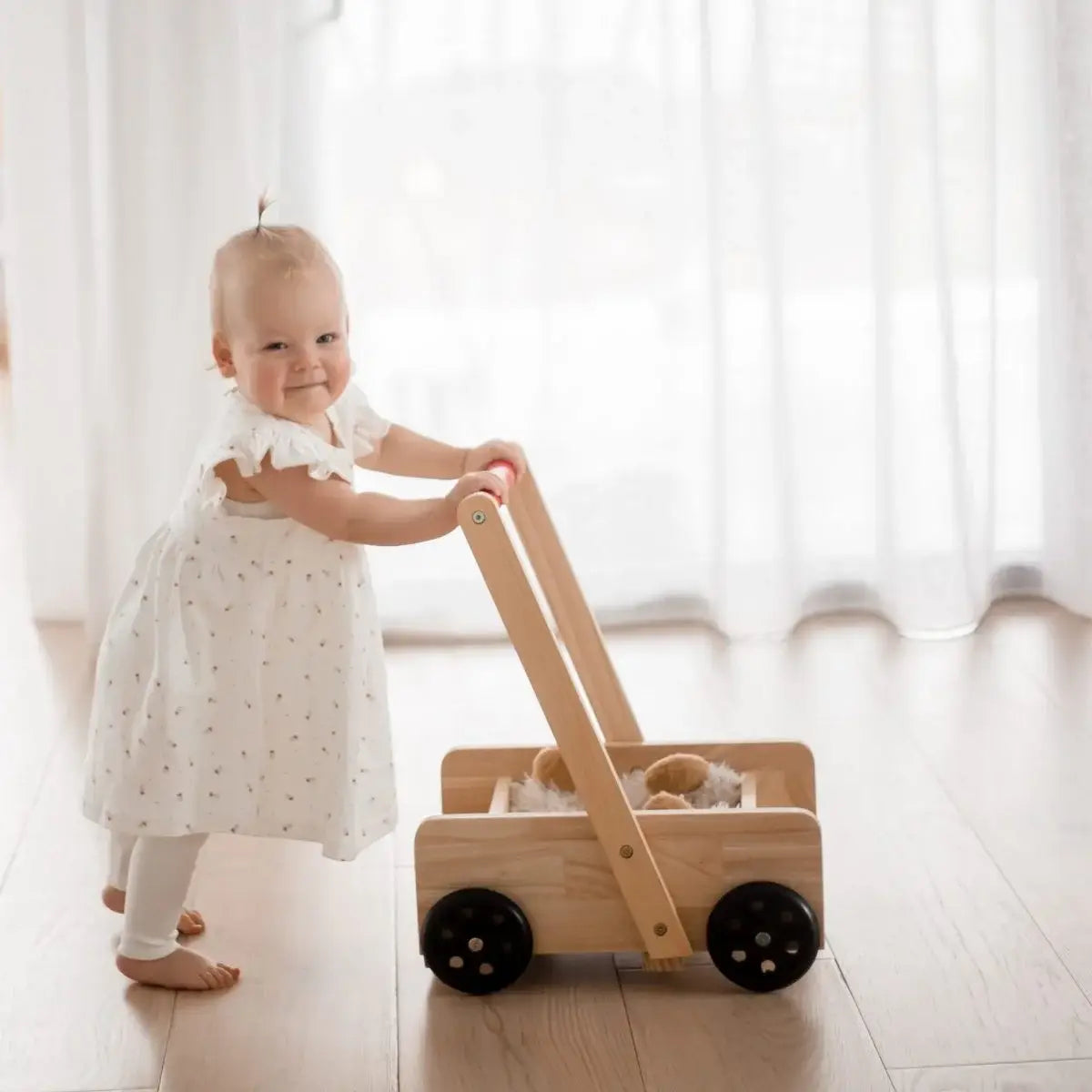 Baby in a white dress pushing a wooden baby walker in a bright room.