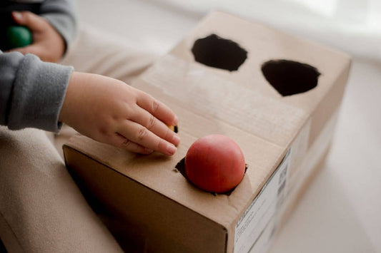 Child's hand pushing colorful wooden balls into a cardboard box with cut-out holes on a light background