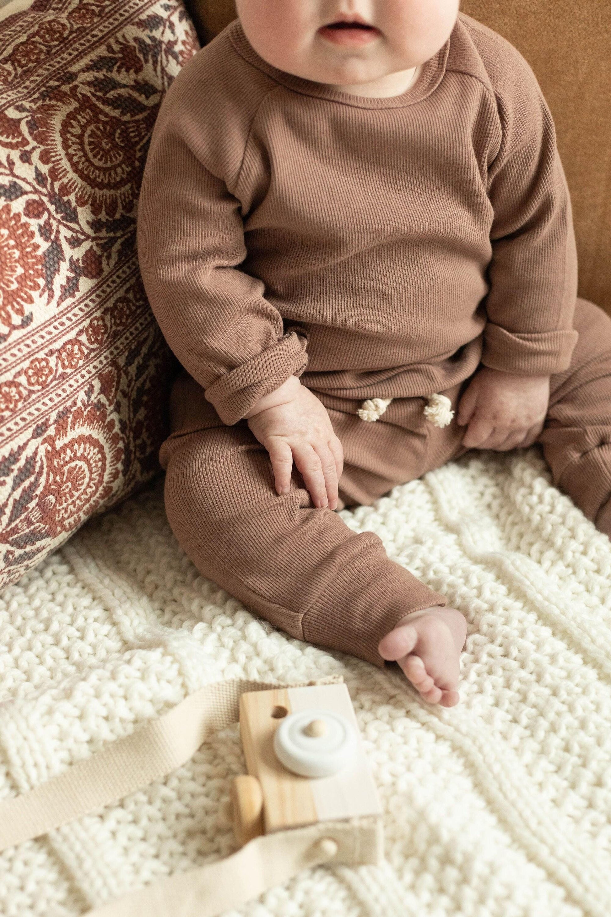 Baby in brown outfit sitting on a textured blanket with a wooden toy.
