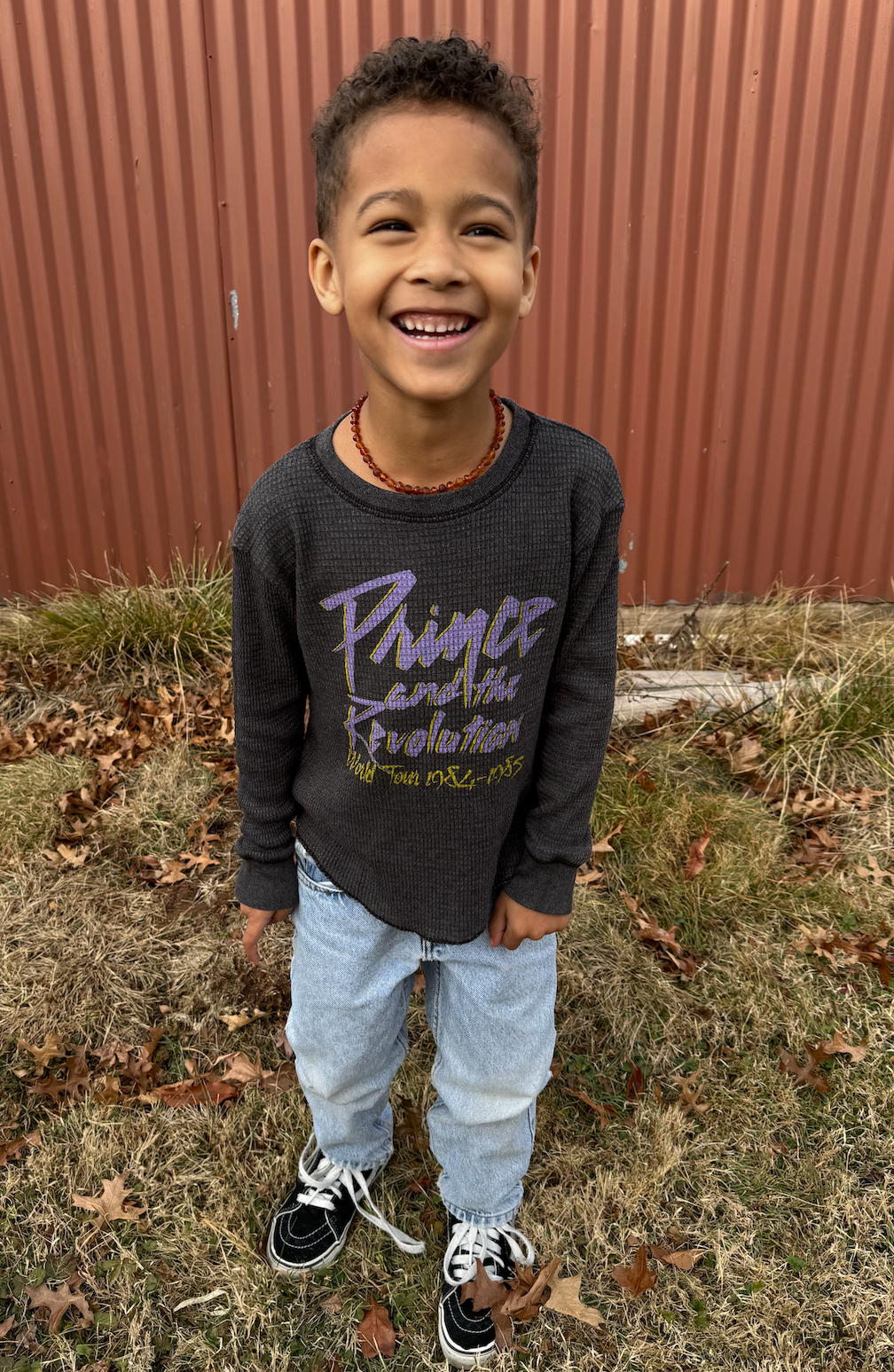 Child wearing a dark gray thermal tee with text, standing outdoors in front of a red metal fence.