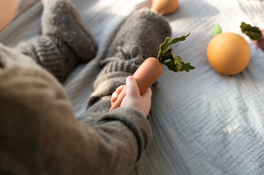 a child sitting on a bed holding a wooden toy carrot with more wooden fruits and vegetabes next to them