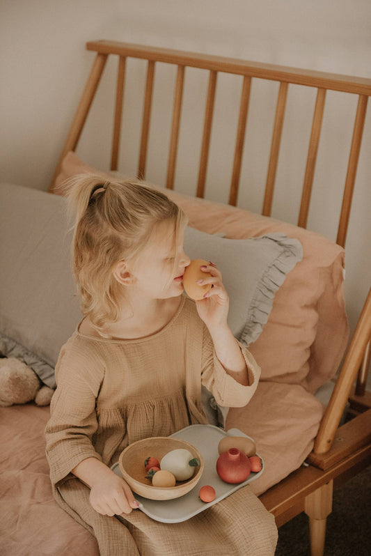a child sitting on a bed playing with wooden fruits on a tray