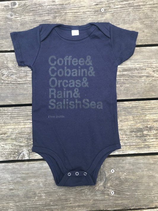 a navy onesie on a wooden table with the words "Coffee& Cobain& Orcas& Rain& Salish Sea" printed on it