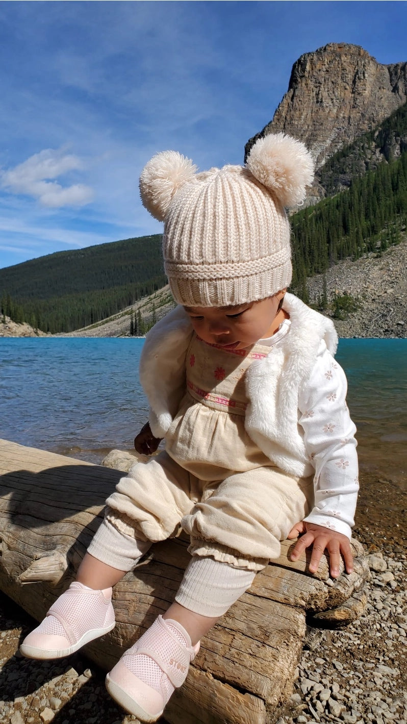 Child in a cream outfit sitting on a log by a lake with mountains in the background wearing natural rubber pink shoes