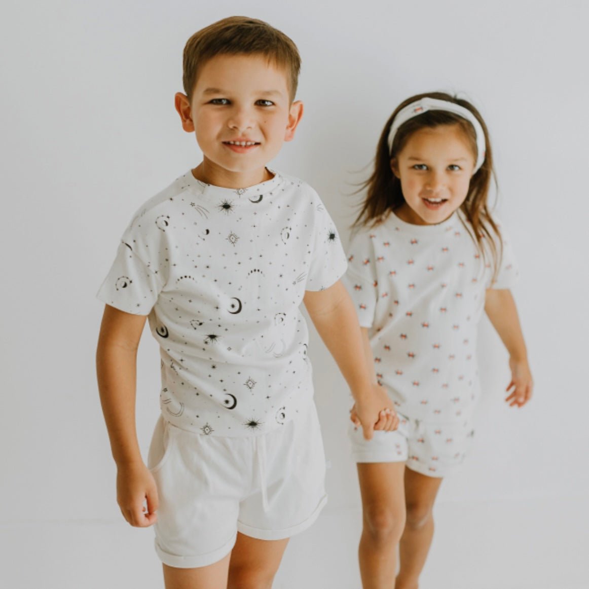 Two children holding hands wearing matching outfits with space-themed designs on a plain background