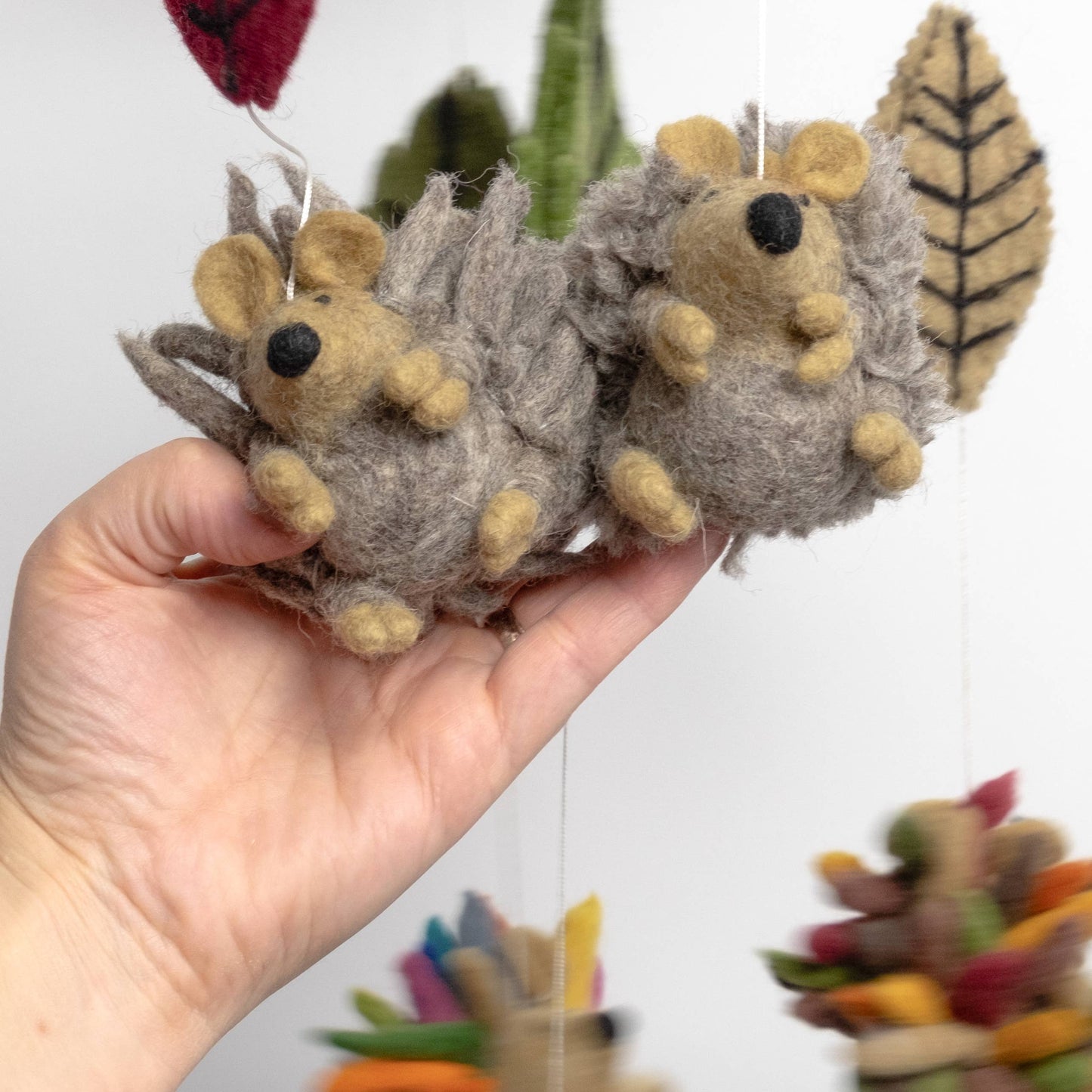 Hand holding two small hedgehog mobile parts with a blurred background of leaves.