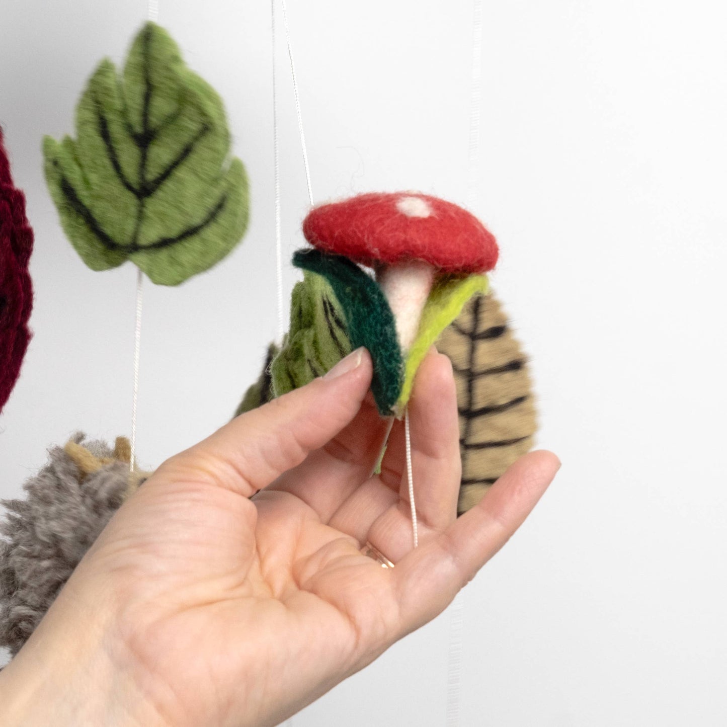 Hand holding a felted mushroom mobile part with green leaves on a white background