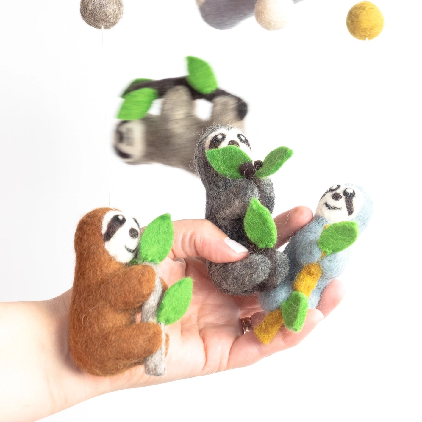Hand holding three small sloth toys hanging from a mobile with green leaves on a white background