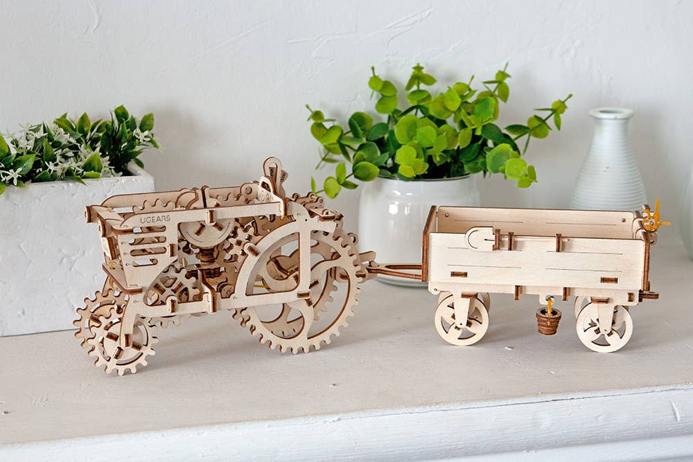 Wooden tractor model with a trailer on a white surface with plants in the background