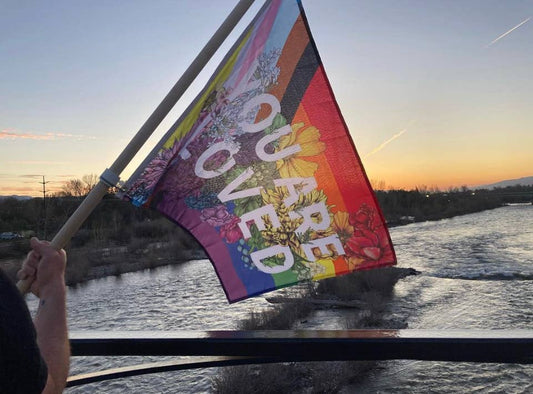 someone holding a flagpole with an intersex inclusive progress pride flag decorated with florals and with the words "you are loved" printed on it hanging from it