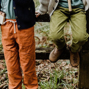 Two children sitting on a fence, one wiring Jackalo's olive green corduroy pants, the other wearing Jackalo's rust corduroy pants.