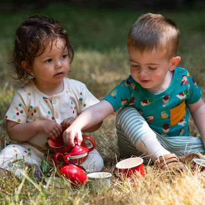 Two babies playing with a tea set in a grass field, one wearing Rainbow Balloons Organic Summer Romper
