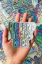 Load image into Gallery viewer, a hand holding a holographic vinyl sticker with the words &quot;Defend Trans Kids&quot;
